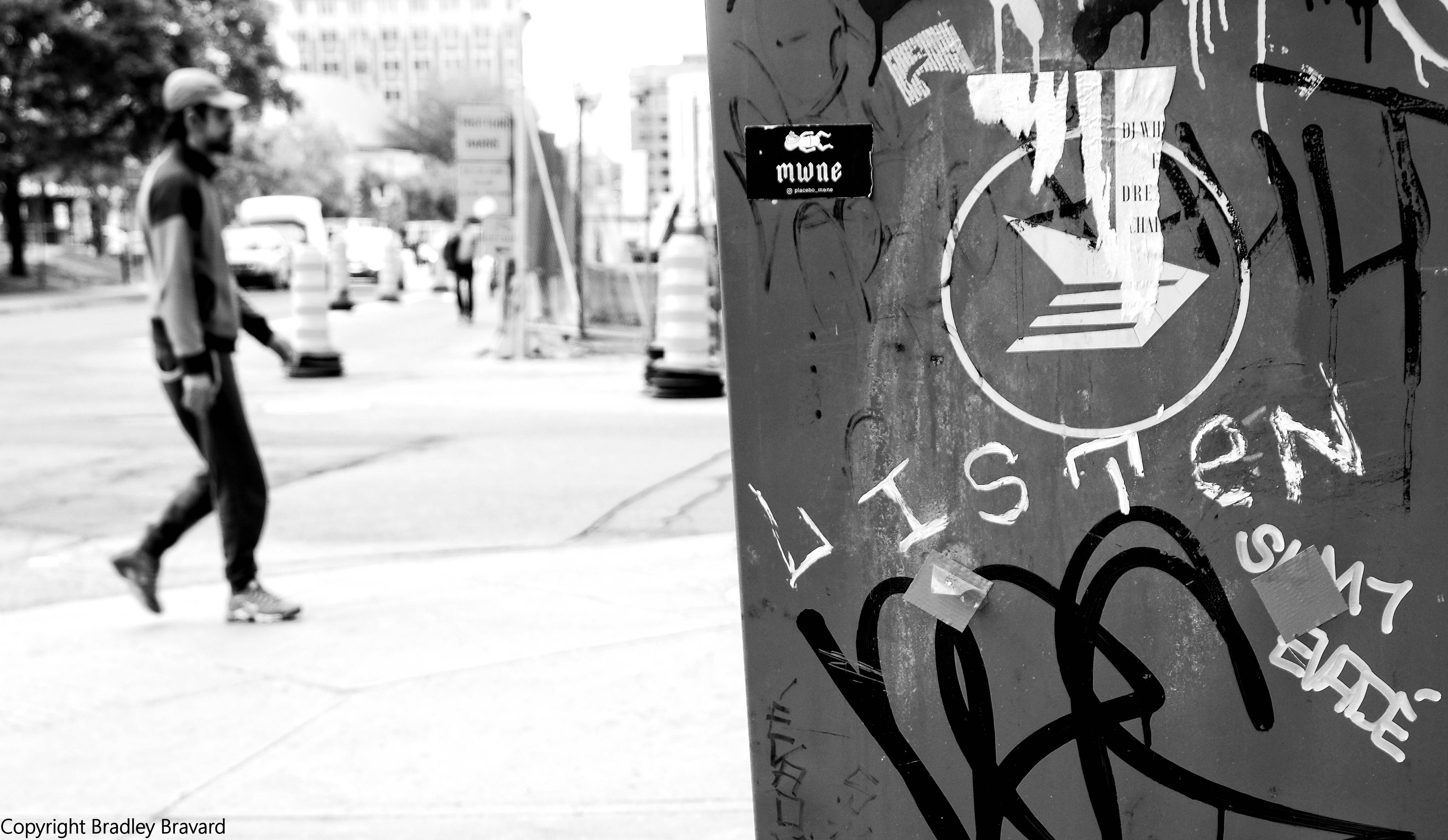 Man walking on sidewalk with graffiti "Listen" on utility post