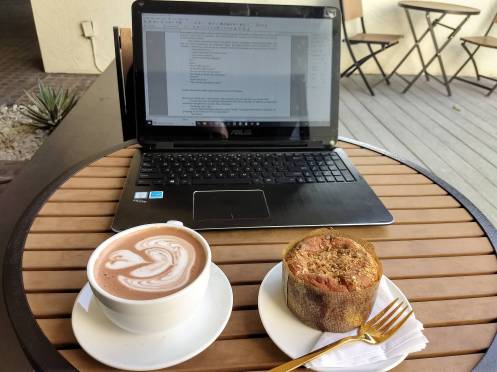 Photo of laptop computer on outdoor table at a coffee shop, with a muffin and a mug of hot chocolate