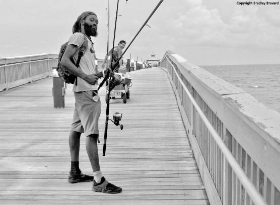 Black and white photo of man carrying fishing poles on pier at Atlantic Ocean coastline
