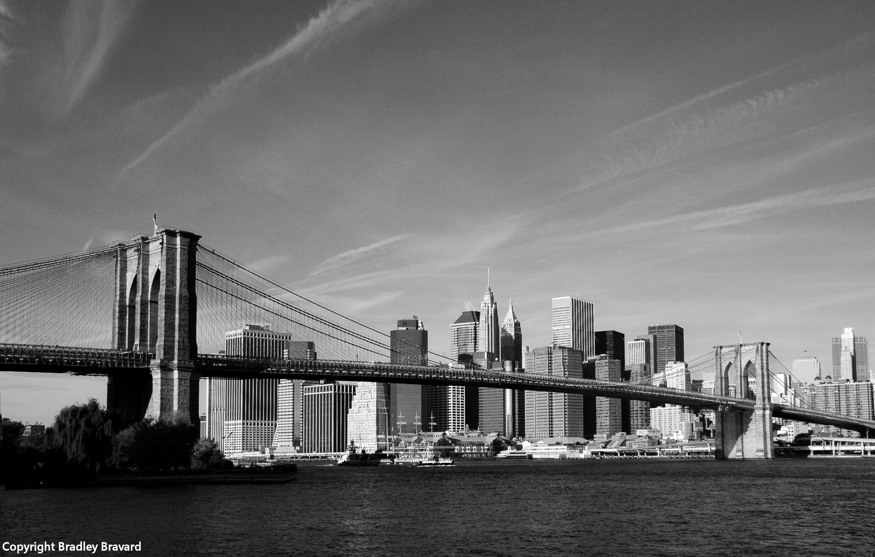 Black and white photo of lower Manhattan skyline behind the Brooklyn Bridge viewed across the East River