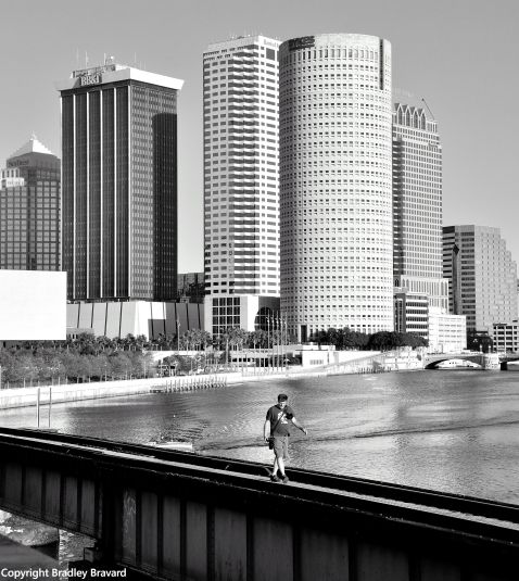 Black and white photo of downtown Tampa skyline, with a person crossing the Hillsborough River on a footbridge in the foreground