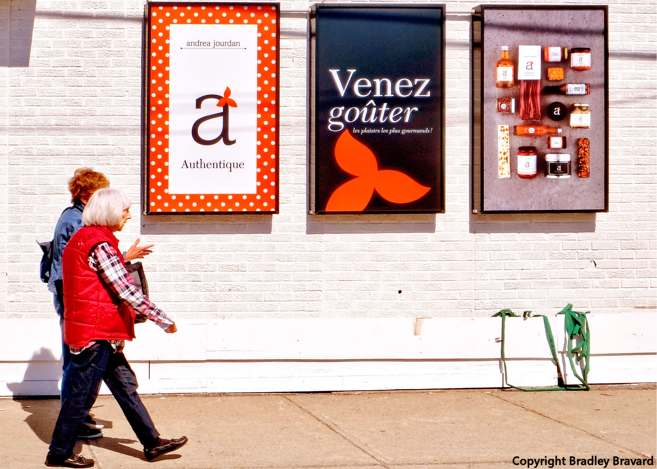 Photo of two women walking on sidewalk next to 3 posters on white brick wall
