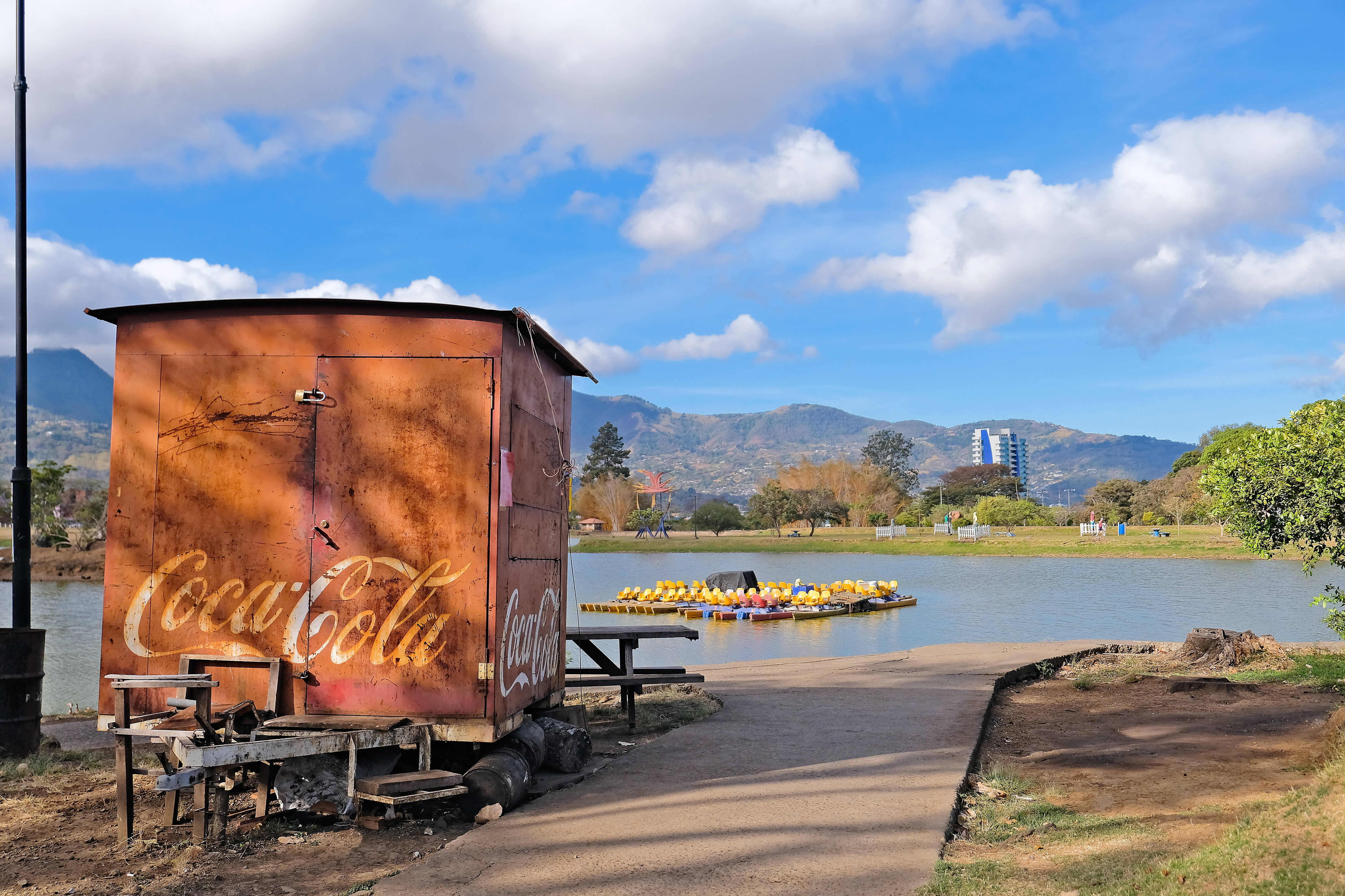 Wooden refreshment stand with Coca Cola logo in front of lake, with mountains and partly cloudy sky in background